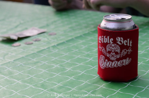 A cold beer can in a koozie with a skull on a picnic tablecloth, with gambling money in the background.